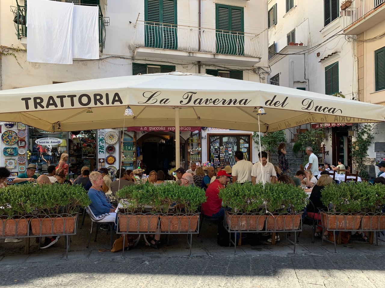 Zona pranzo all'aperto presso La Taverna del Duca ad Amalfi, caratterizzata dalla tradizionale atmosfera mediterranea con persiane verdi e accenti floreali.